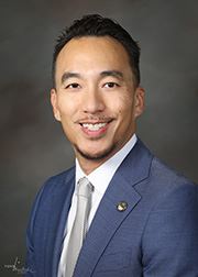 Man in gray suit and blue tie posing outdoors in front of greenery.